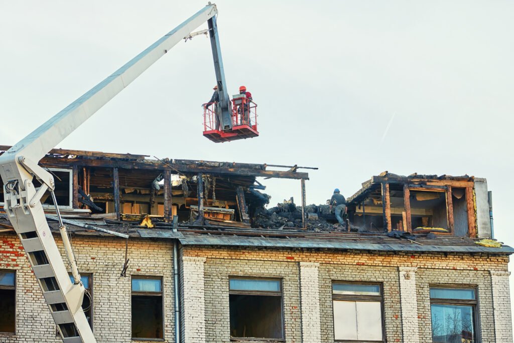 Unrecognizable workers in crane cradle dismantling charred roof after fire, aftermath of an emergency. Men cleanup and restoration building roof after fire.