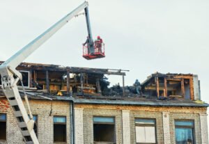 Unrecognizable workers in crane cradle dismantling charred roof after fire, aftermath of an emergency. Men cleanup and restoration building roof after fire.